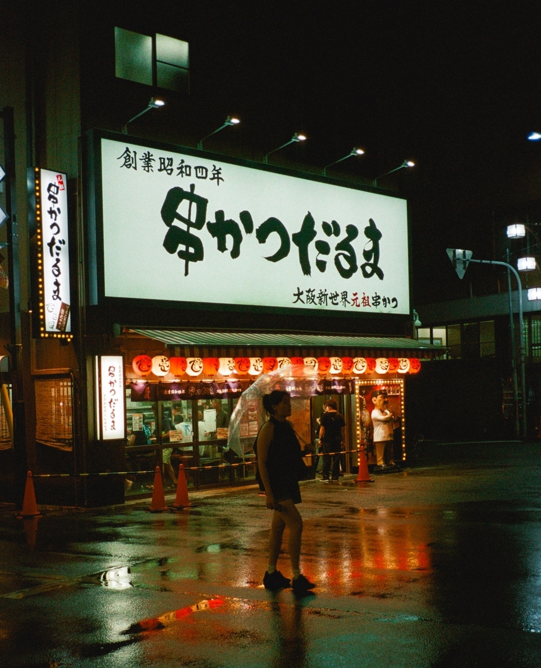 Woman with umbrella stands on a wet street in front of a Japanese restaurant at night, with signage and red lanterns.