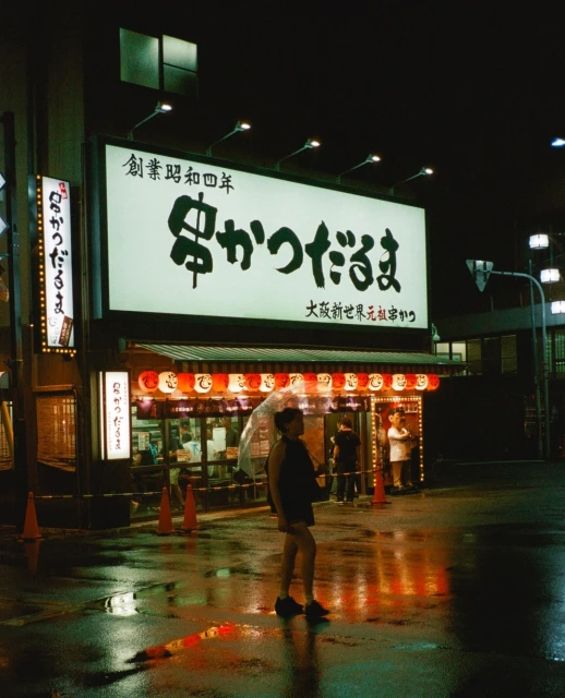 Woman with umbrella stands on a wet street in front of a Japanese restaurant at night, with signage and red lanterns.