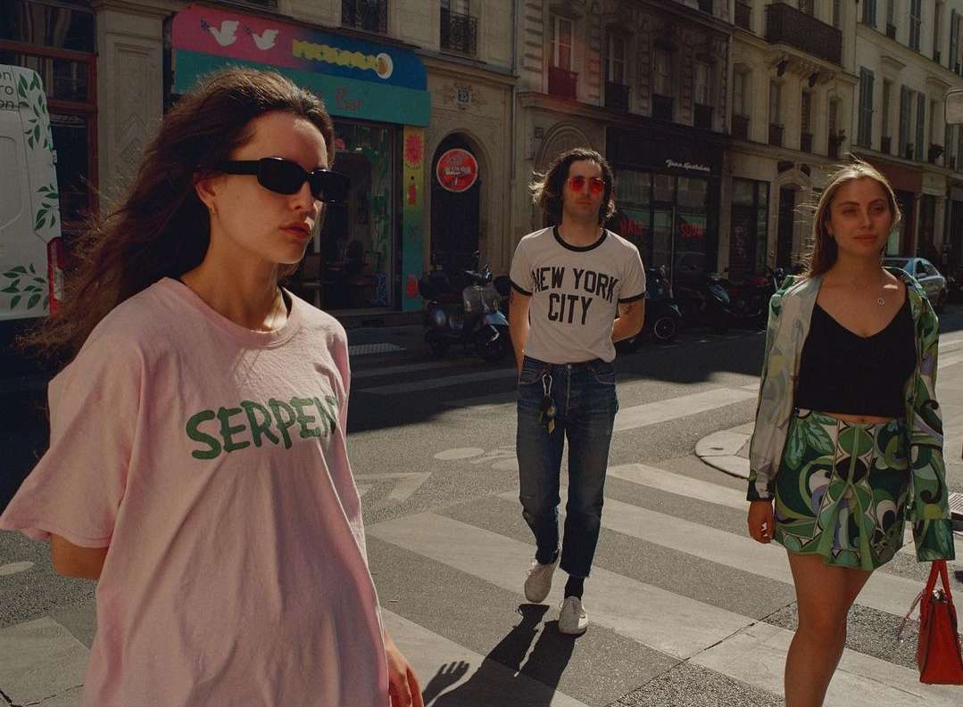 Three people walking on a zebra crossing in a European city. A woman in a pink t-shirt is in the foreground, with two people behind her.