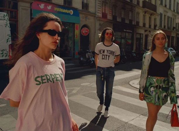 Three people walking on a zebra crossing in a European city. A woman in a pink t-shirt is in the foreground, with two people behind her.