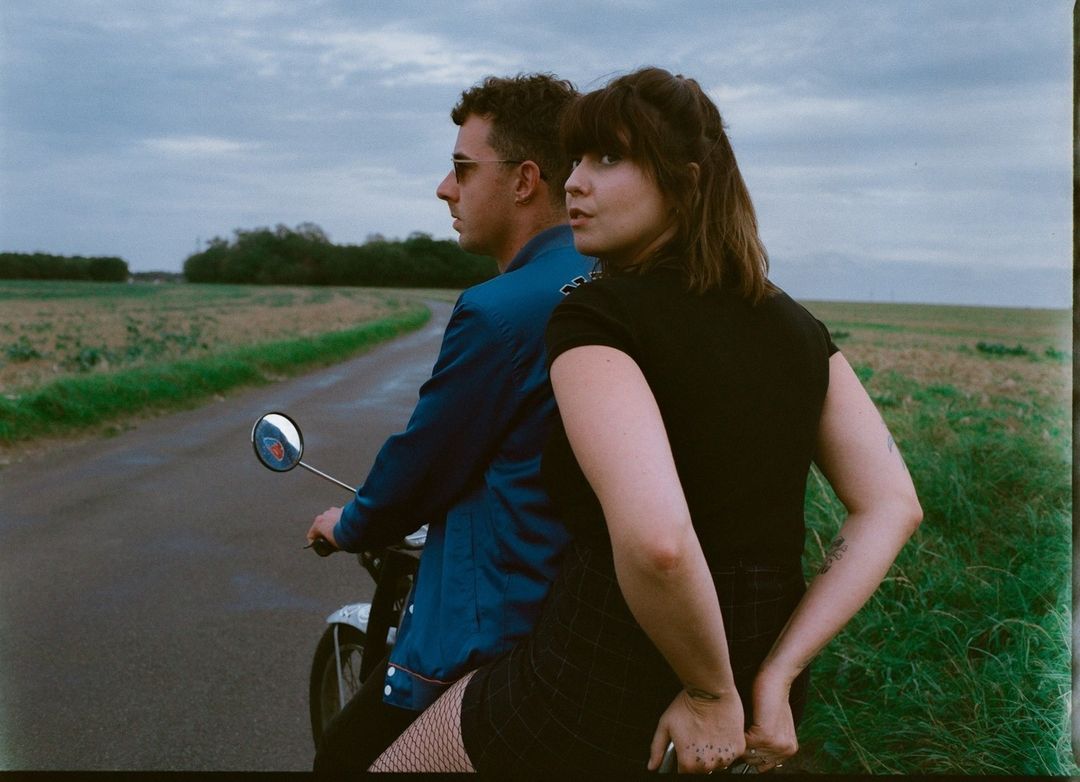 A couple rides a scooter on a rural road under a cloudy sky. The man drives while the woman sits behind him, looking off to the side.