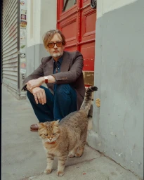 Man crouching outside near a tabby cat in front of a red doorway in Paris