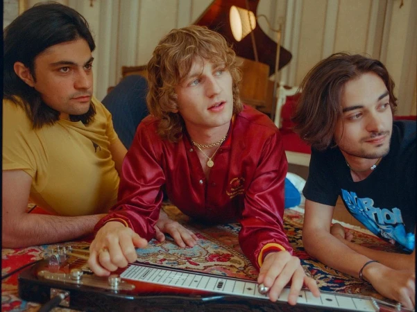 Three young men posing together indoors, with the central figure playing a lap steel guitar on a patterned carpet floor.