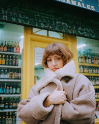 Woman with short brown hair wearing a shearling-lined beige coat standing outside a Parisian storefront with shelves of bottled goods.