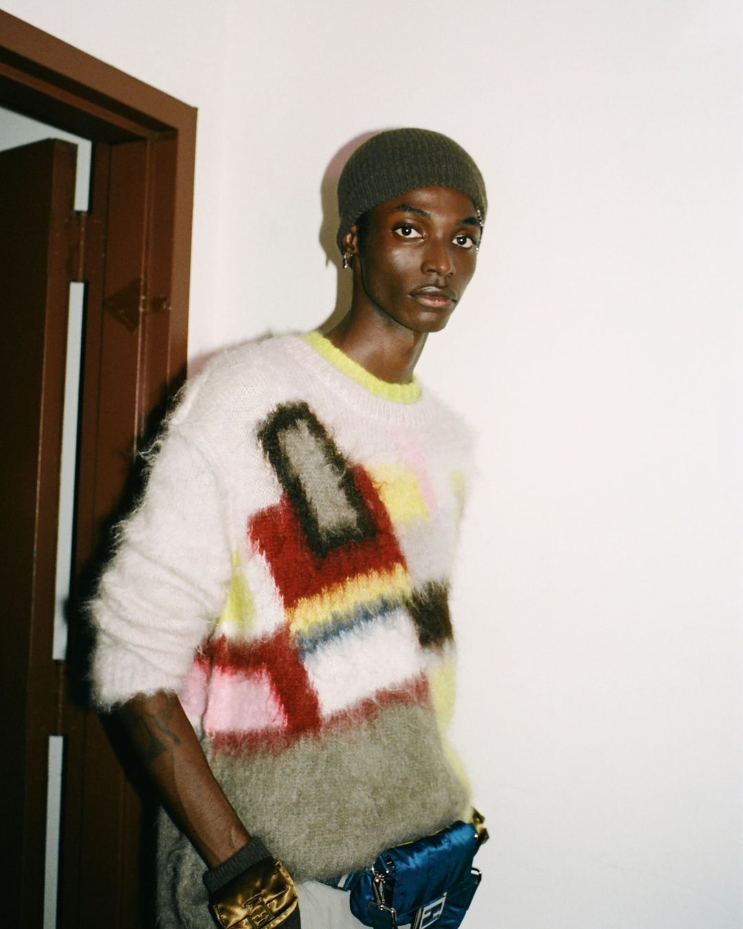 A young Black man is posing indoors wearing a colorful sweater, olive-green beanie, and a blue purse; the room has white walls.