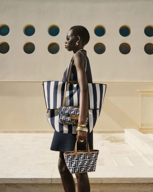 Fashion shot of a black woman walking sideways wearing a blue dress, sunglasses, gold jewelry, and several Fendi branded handbags.