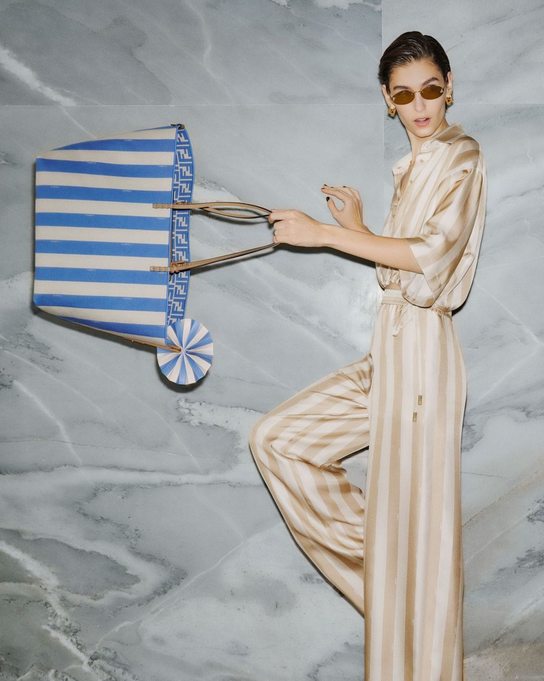 Fashion shot featuring a woman in a striped outfit holding a striped bag in front of a marble wall, styled with tan sunglasses.
