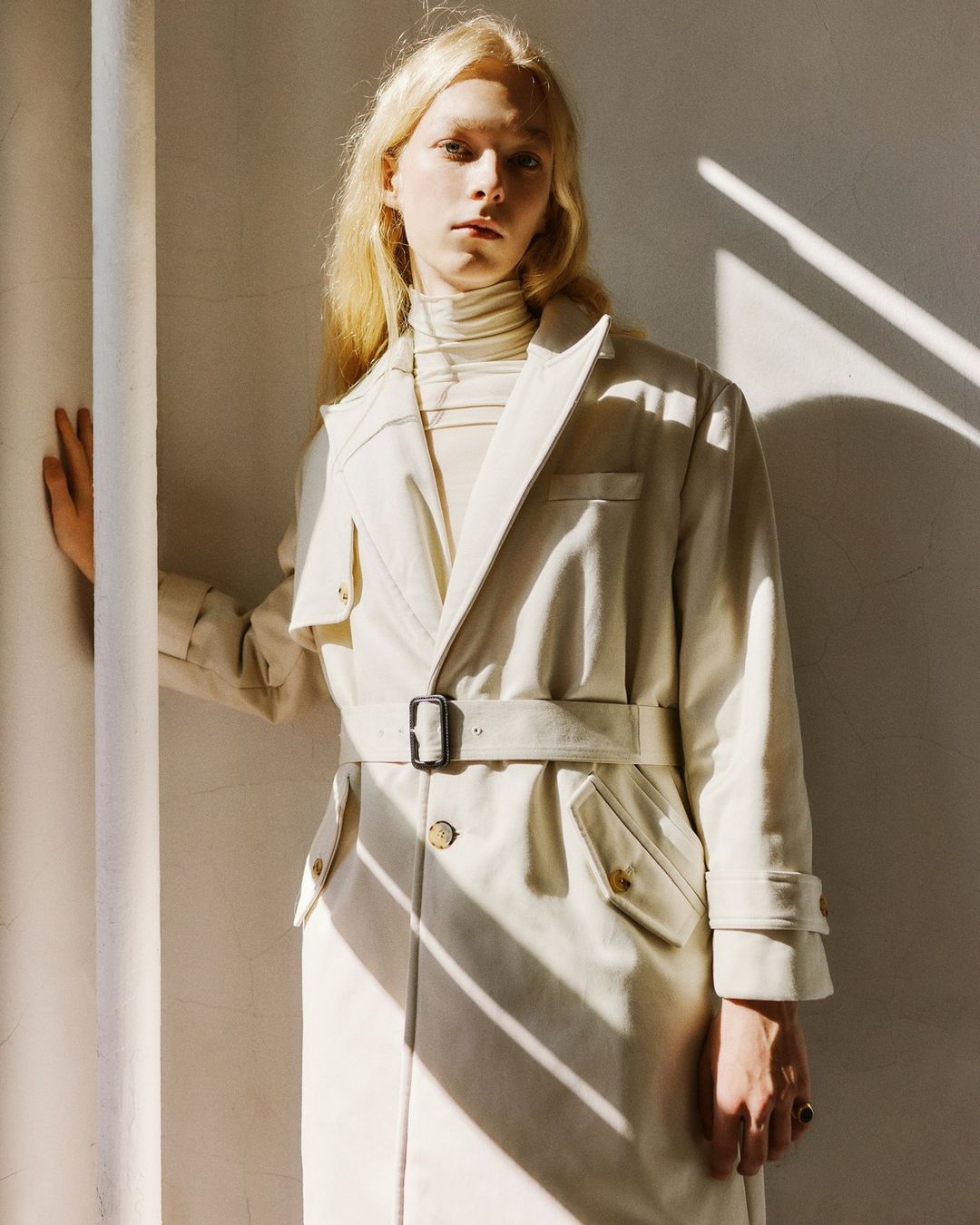 Model with blonde hair wearing a cream belted trench coat and turtleneck standing in harsh sunlight by a pillar