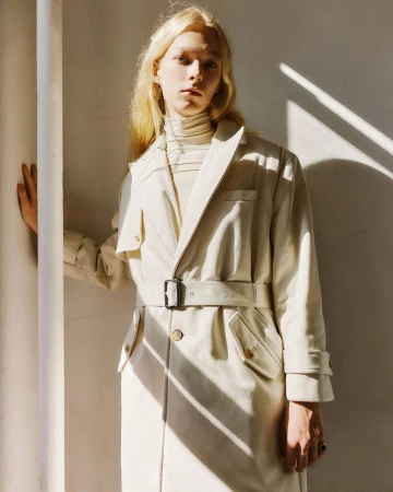Model with blonde hair wearing a cream belted trench coat and turtleneck standing in harsh sunlight by a pillar