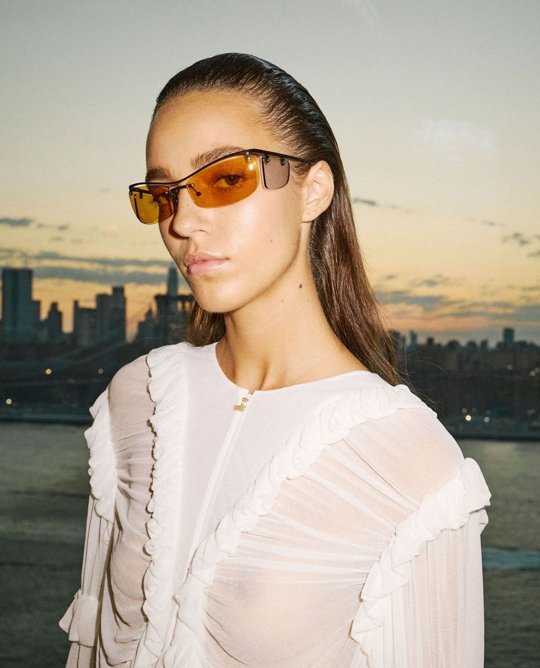 Portrait of a woman wearing sunglasses and a ruffled white top, with a cityscape and river in the background at sunset.