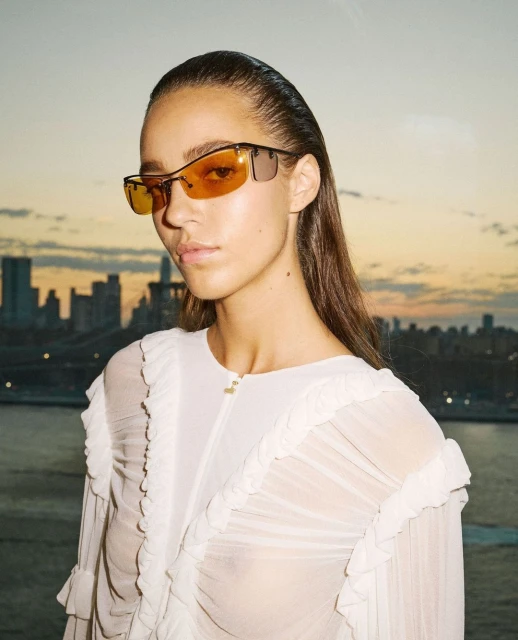 Portrait of a woman wearing sunglasses and a ruffled white top, with a cityscape and river in the background at sunset.