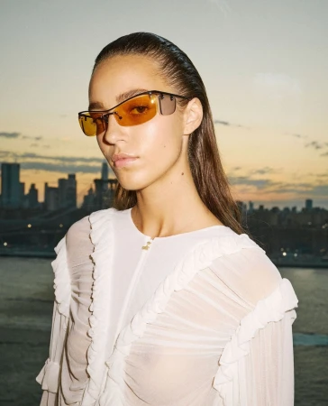 Portrait of a woman wearing sunglasses and a ruffled white top, with a cityscape and river in the background at sunset.