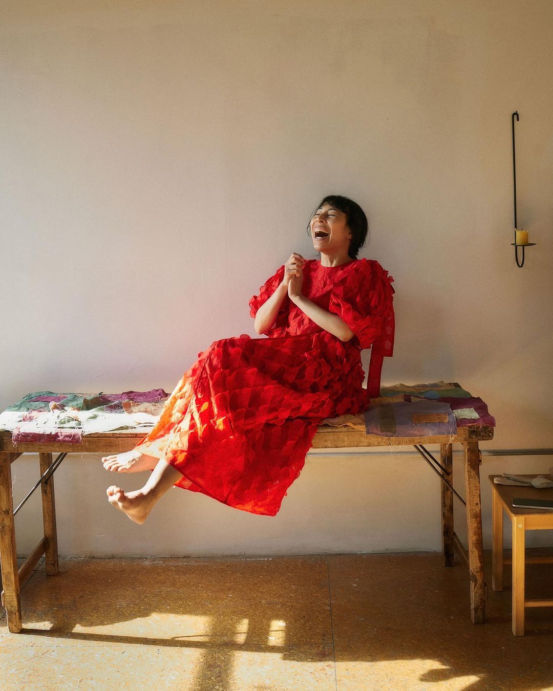A woman in a red dress sits on a rustic table, joyfully laughing with hands clasped together in a bright, sunny room.