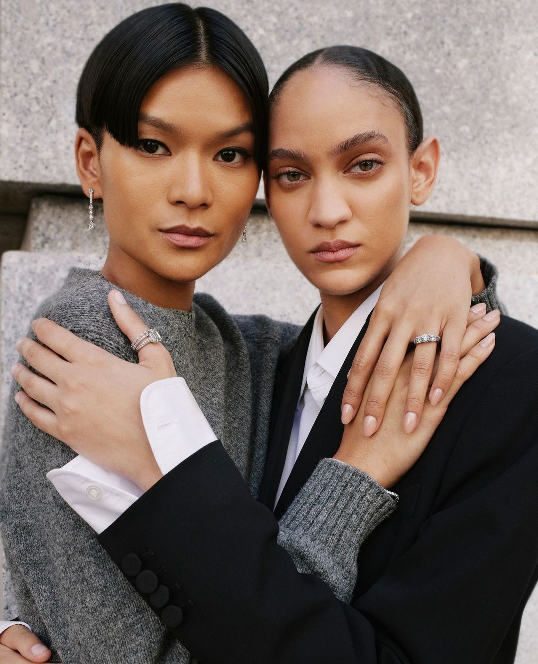 Close-up shot of two adult women with neutral expressions, one in a gray sweater and the other in a black blazer, embracing indoors.