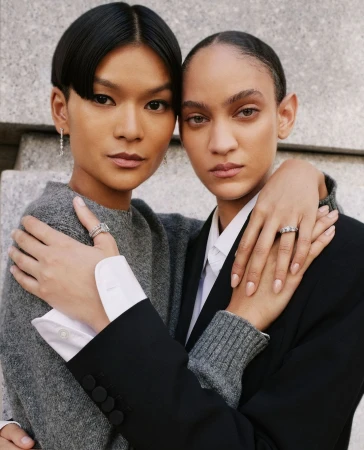 Close-up shot of two adult women with neutral expressions, one in a gray sweater and the other in a black blazer, embracing indoors.