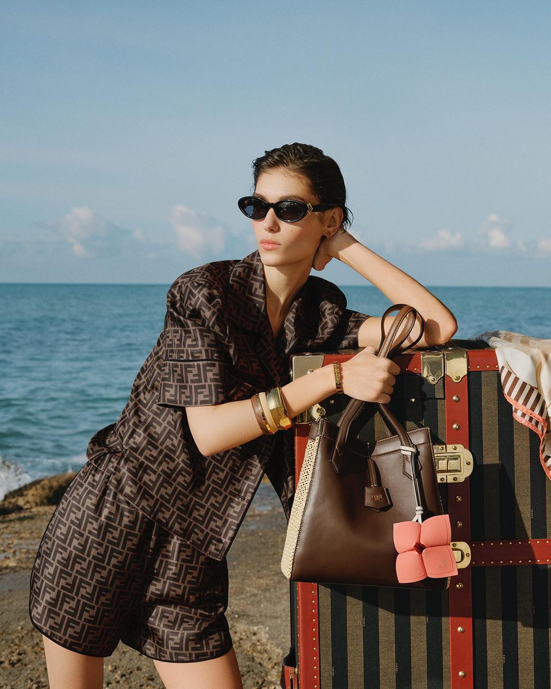 Woman in Fendi patterned outfit, sunglasses, and gold bracelets poses next to a vintage steamer trunk on a beach with ocean backdrop.