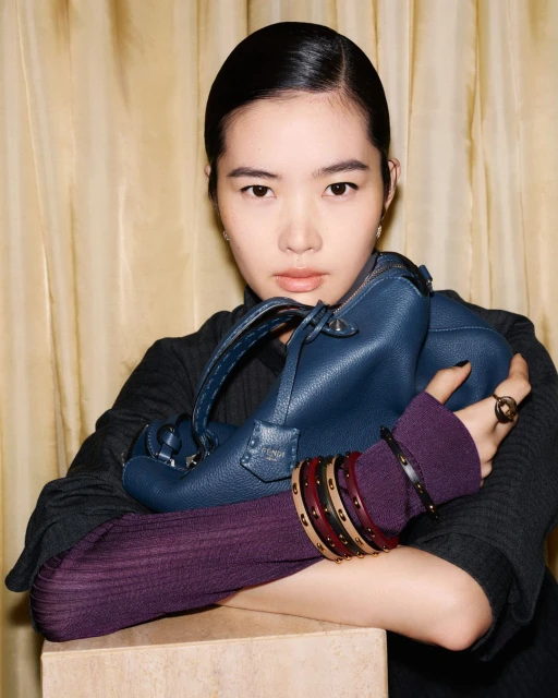 Young Asian woman poses indoors with a textured blue handbag, wearing layered sleeve cuffs and rings, with a neutral backdrop and soft lighting.