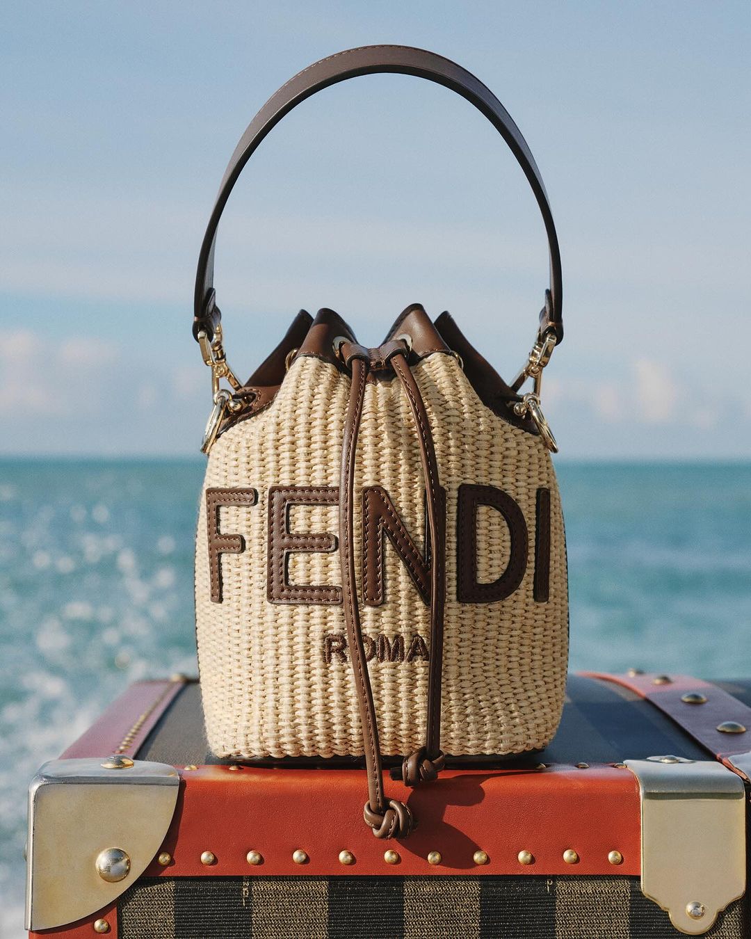 Wicker Fendi Roma bucket bag with leather trim on top of vintage luggage against a sea and sky background, fashion advertisement.