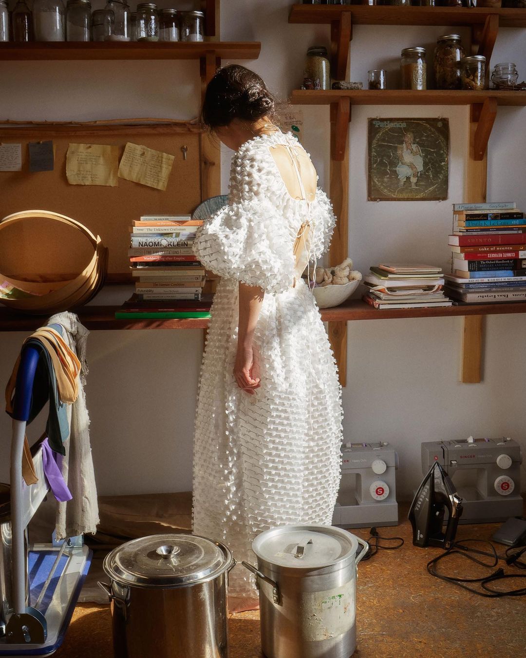 Woman in white textured dress stands in sunlit domestic setting with shelves, pots, and sewing machines visible indoors.
