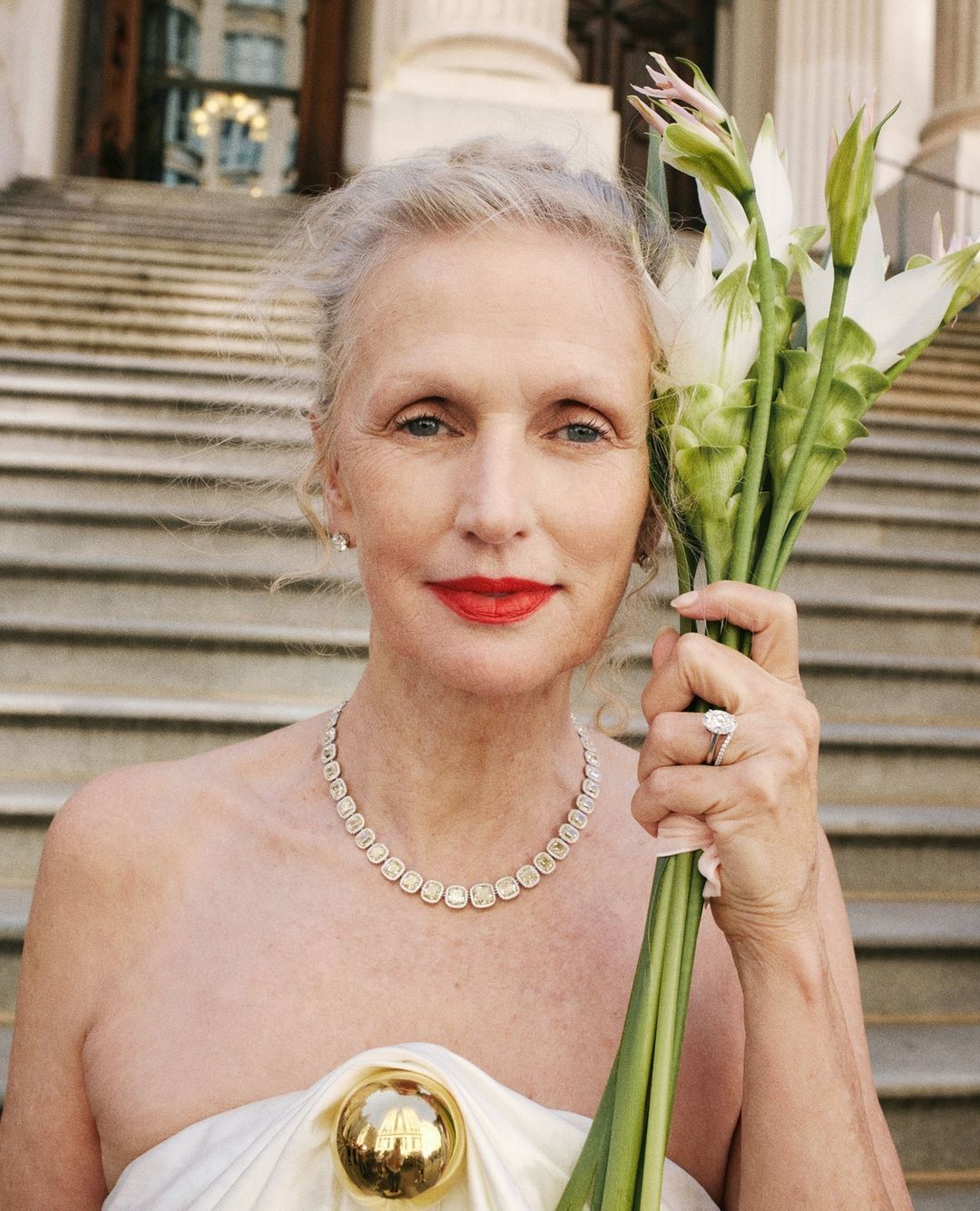 Elegant older woman in strapless dress, accessorized with jewelry and holding flowers, poses against a backdrop of classic architecture.