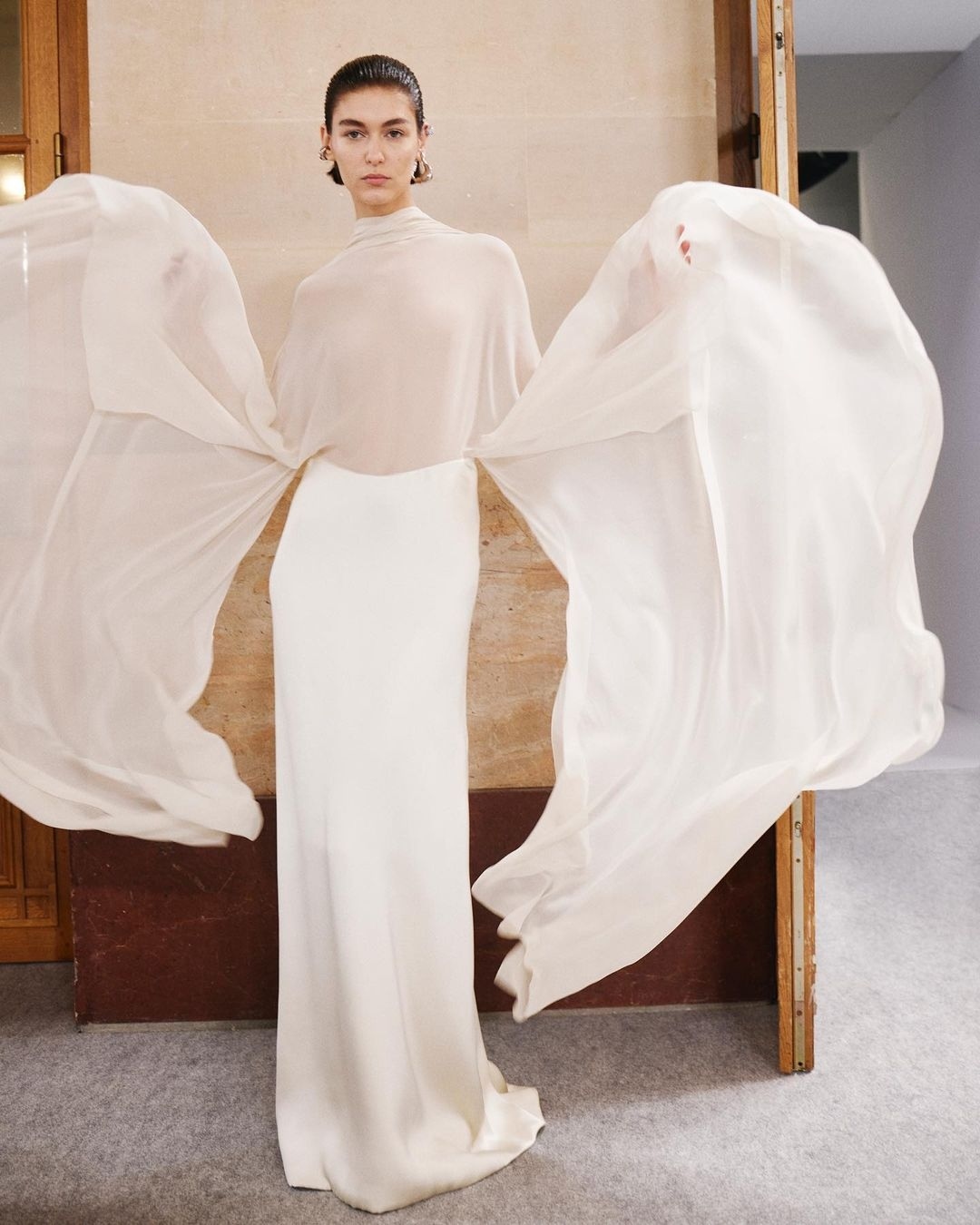 Elegant woman in a flowing white gown with sheer sleeves poses in a neutral indoor setting for a fashion editorial photograph.