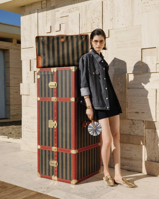 A young woman stands beside a striped trunk and luggage set in an outdoor setting, wearing a dark jacket, dress, and tan loafers.