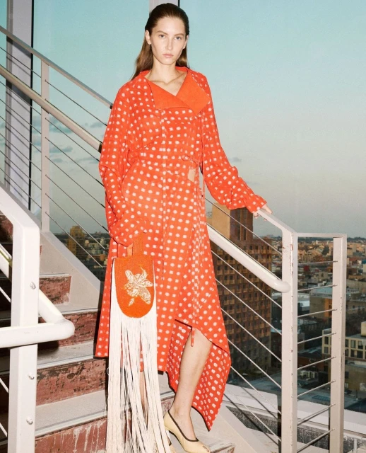 Woman in orange polka-dot dress poses on staircase with beaded handbag. Urban background with visible cityscape in daylight.