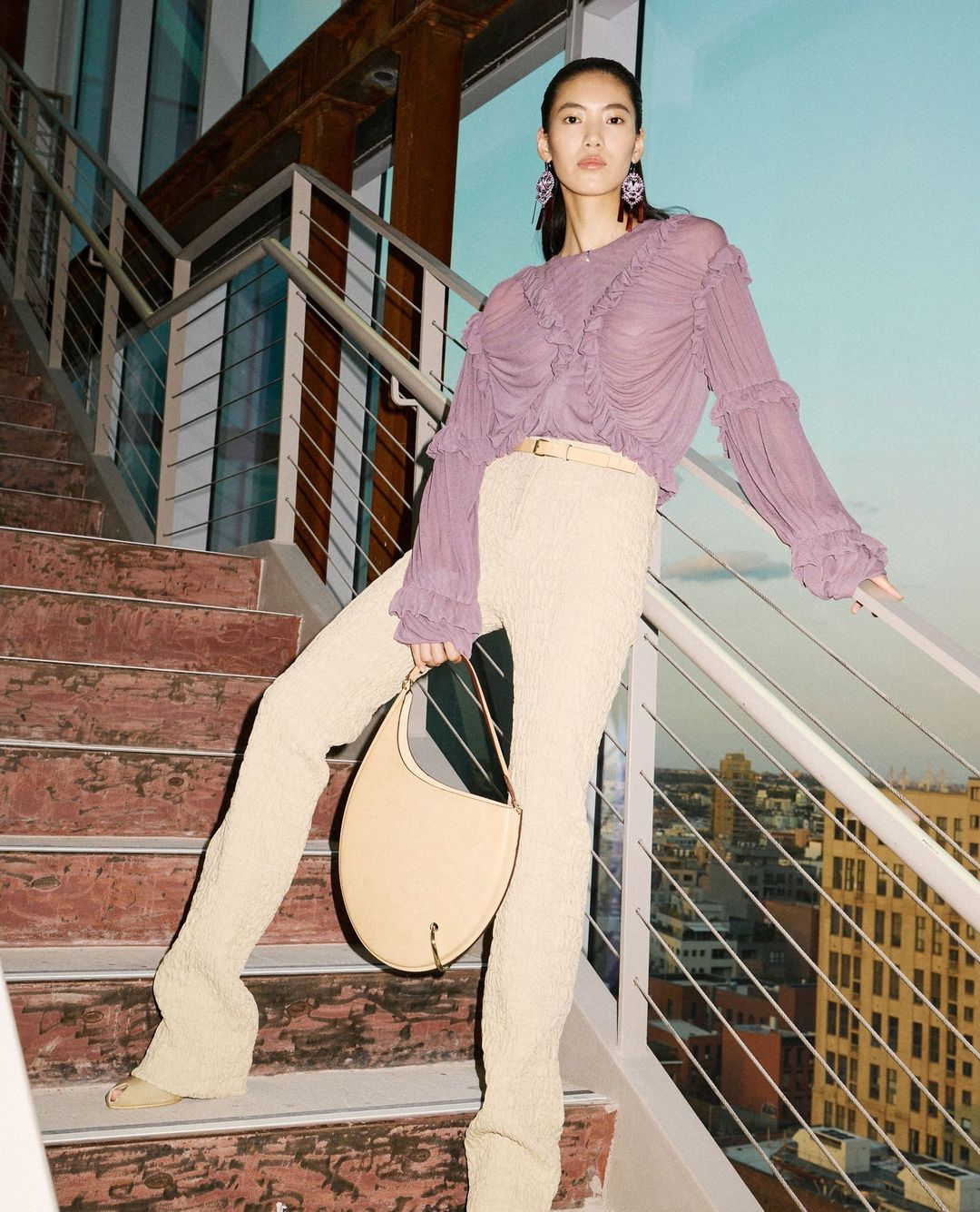 Fashionable young Asian woman poses on stairs with modern architecture, wearing lavender top, light pants, carrying handbag, earrings.