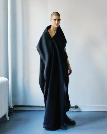 A person with a shaved head wearing a long black sculptural garment standing against a plain wall.