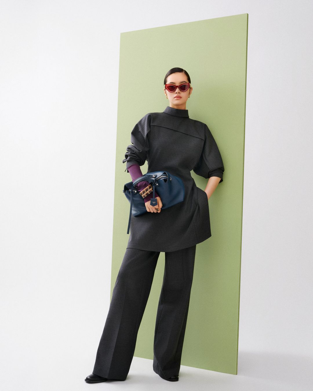 A woman in an elegant gray pantsuit with a blue bag and red sunglasses poses in front of a light green panel in a fashion shot.