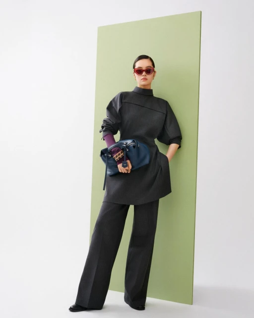 A woman in an elegant gray pantsuit with a blue bag and red sunglasses poses in front of a light green panel in a fashion shot.