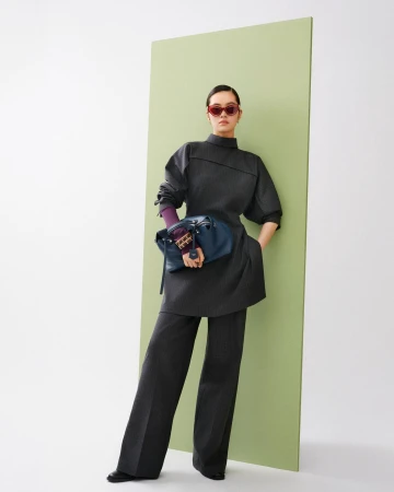 A woman in an elegant gray pantsuit with a blue bag and red sunglasses poses in front of a light green panel in a fashion shot.