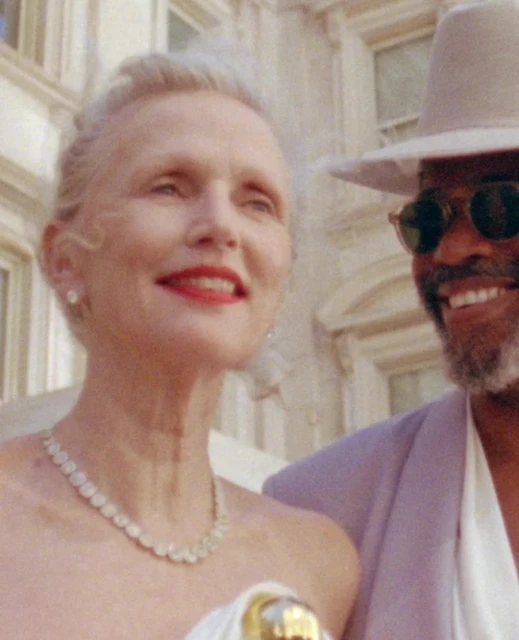 Close-up of an elegant adult woman with fair skin and gray hair wearing a necklace, standing next to an adult male with a hat.