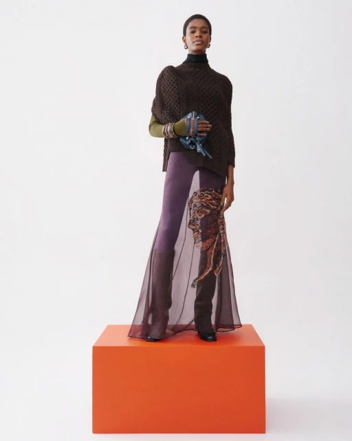 A stylish woman stands on an orange cube, showcasing a brown poncho sweater, purple pants, black boots, and a unique blue bag.