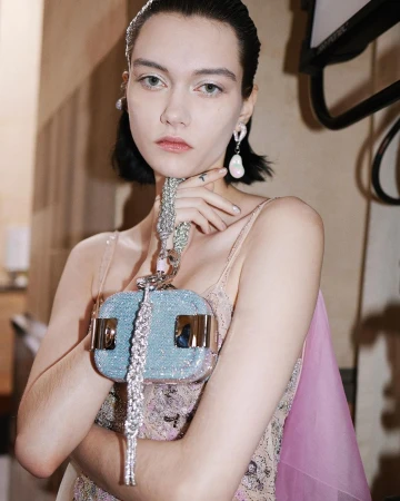 Model with dark chin-length hair wearing drop pearl earrings and holding a shimmering light blue textured handbag by its silver chain strap.