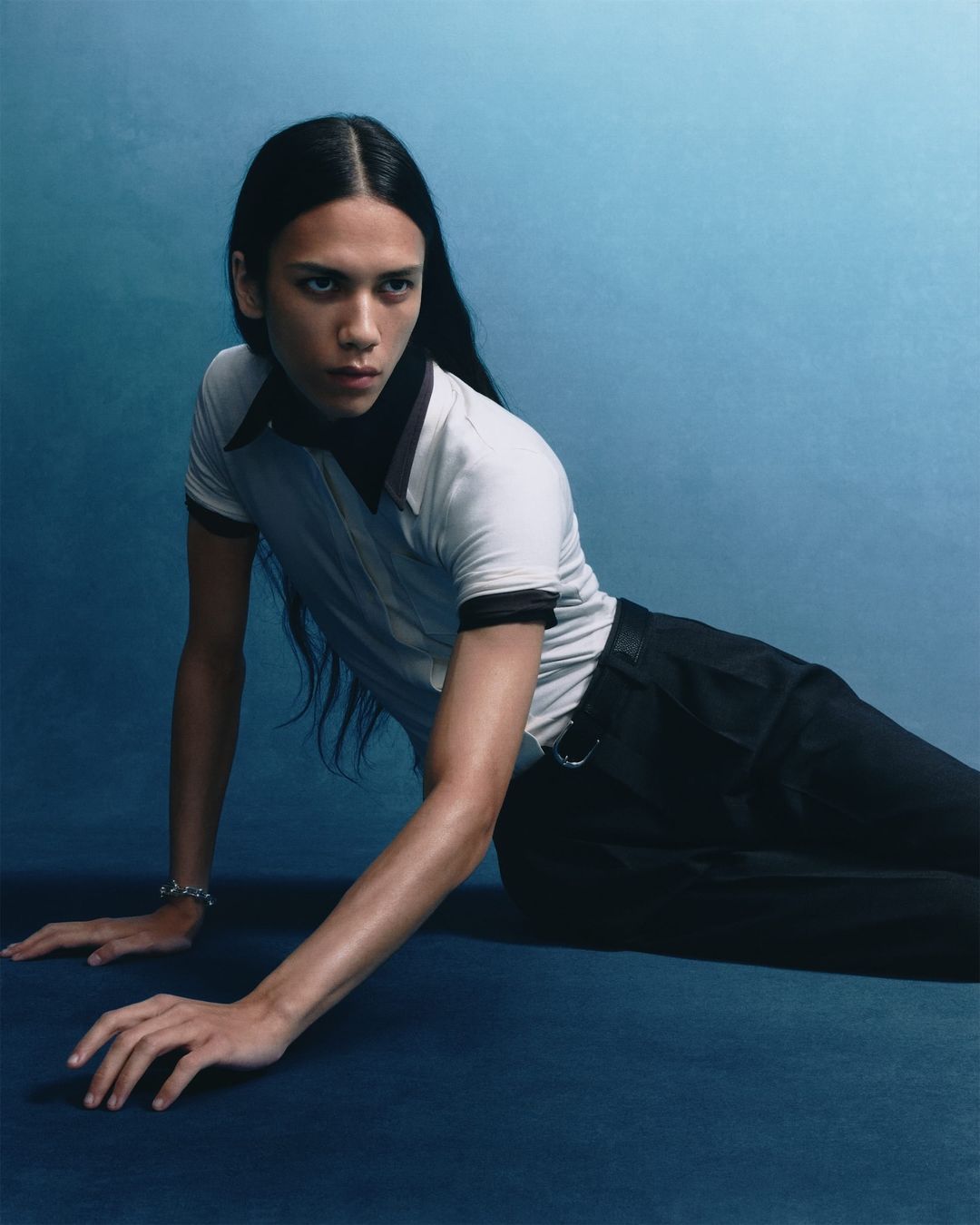 Portrait of a young adult with long dark hair wearing a white top with black trim and black pants against a gradient blue background.