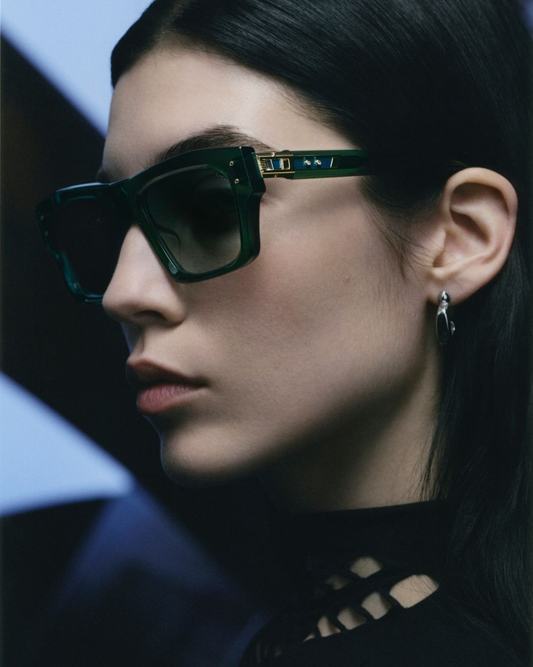 A woman poses in a close-up studio shot wearing green, square-framed sunglasses and silver hoop earrings, against a dark background.