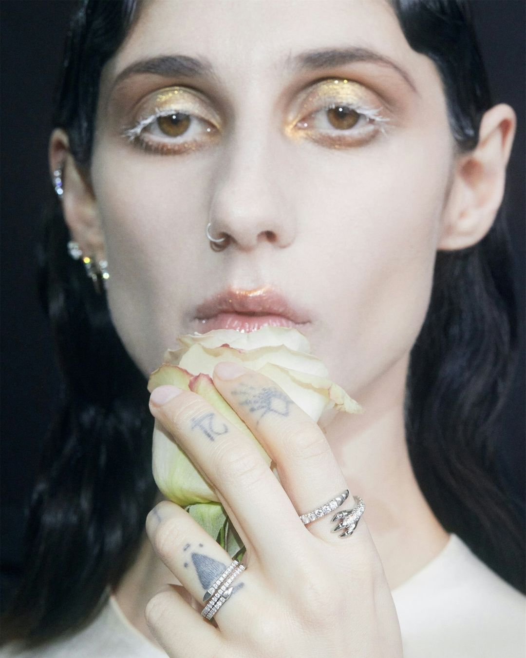 Close-up portrait of a woman with gold eye makeup, a nose ring, and finger tattoos holding a white rose in a dark studio setting.