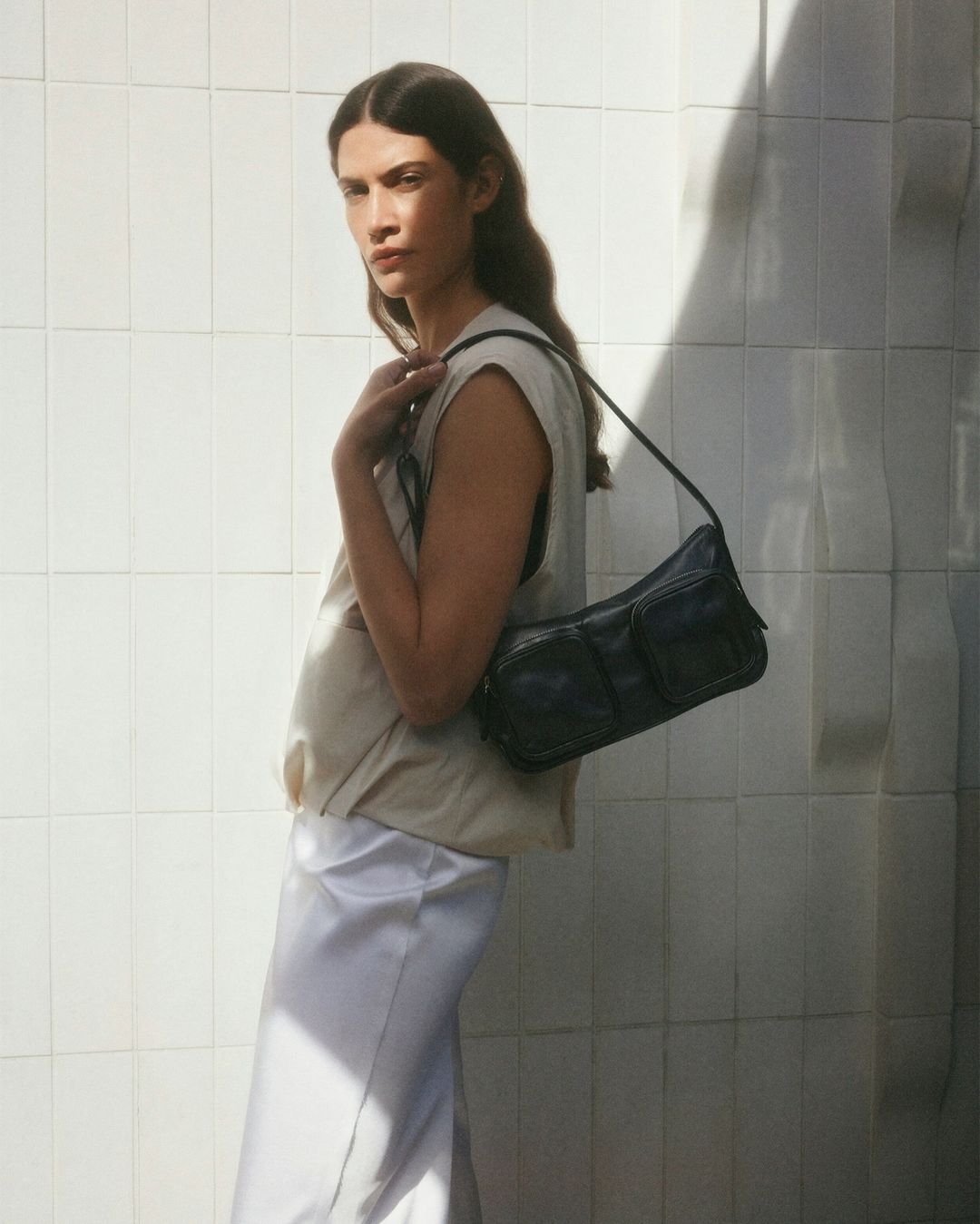 Woman poses with a black shoulder bag in front of a white tile wall, illuminated by sunlight creating shadows.