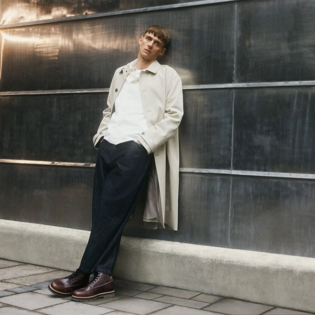 A fashionable adult man leans against a wall wearing a light beige trench coat, white shirt, dark blue pants, and brown leather dress shoes.
