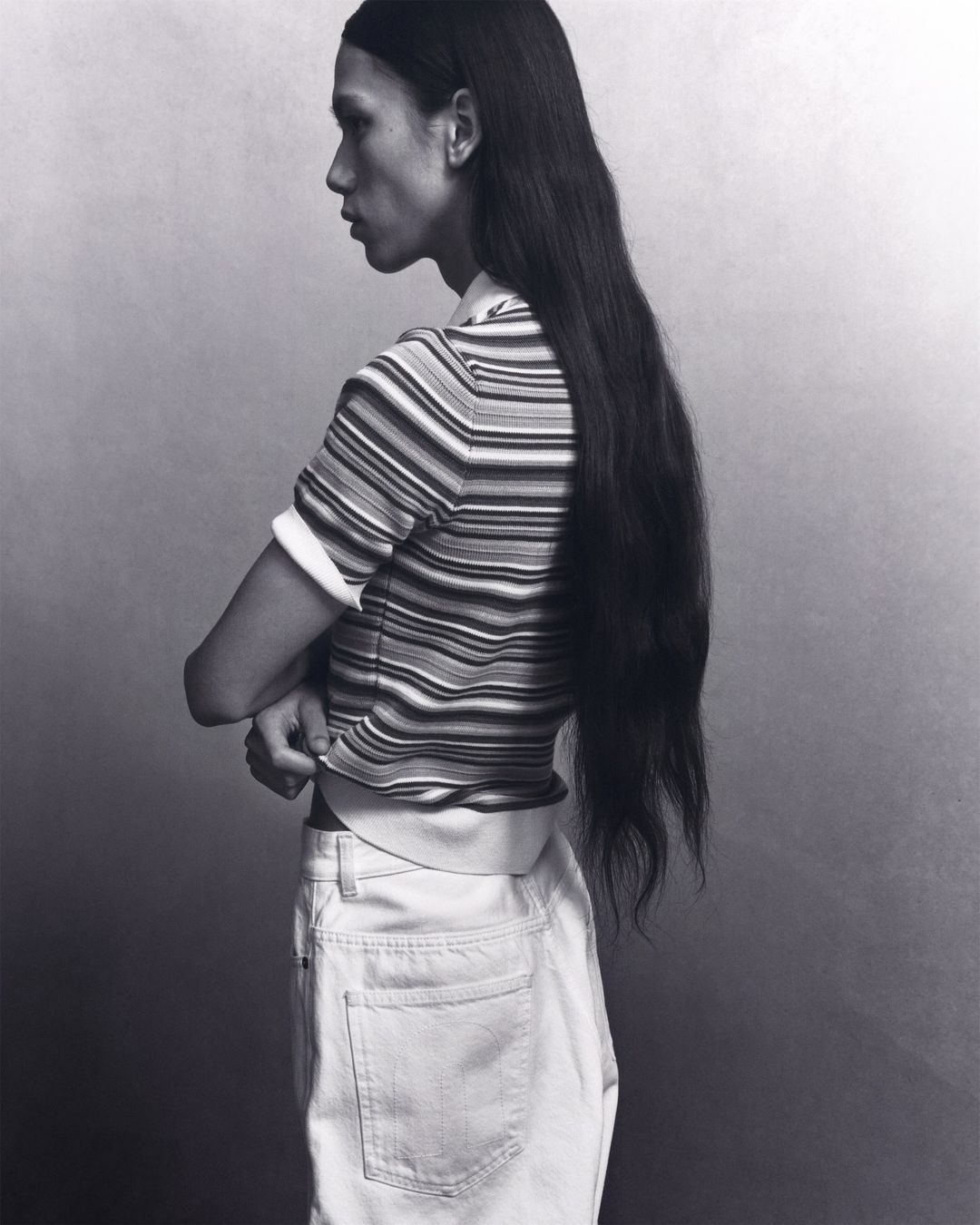 Profile view of a model with long hair wearing a striped shirt and light jeans, set against a soft grey gradient background.