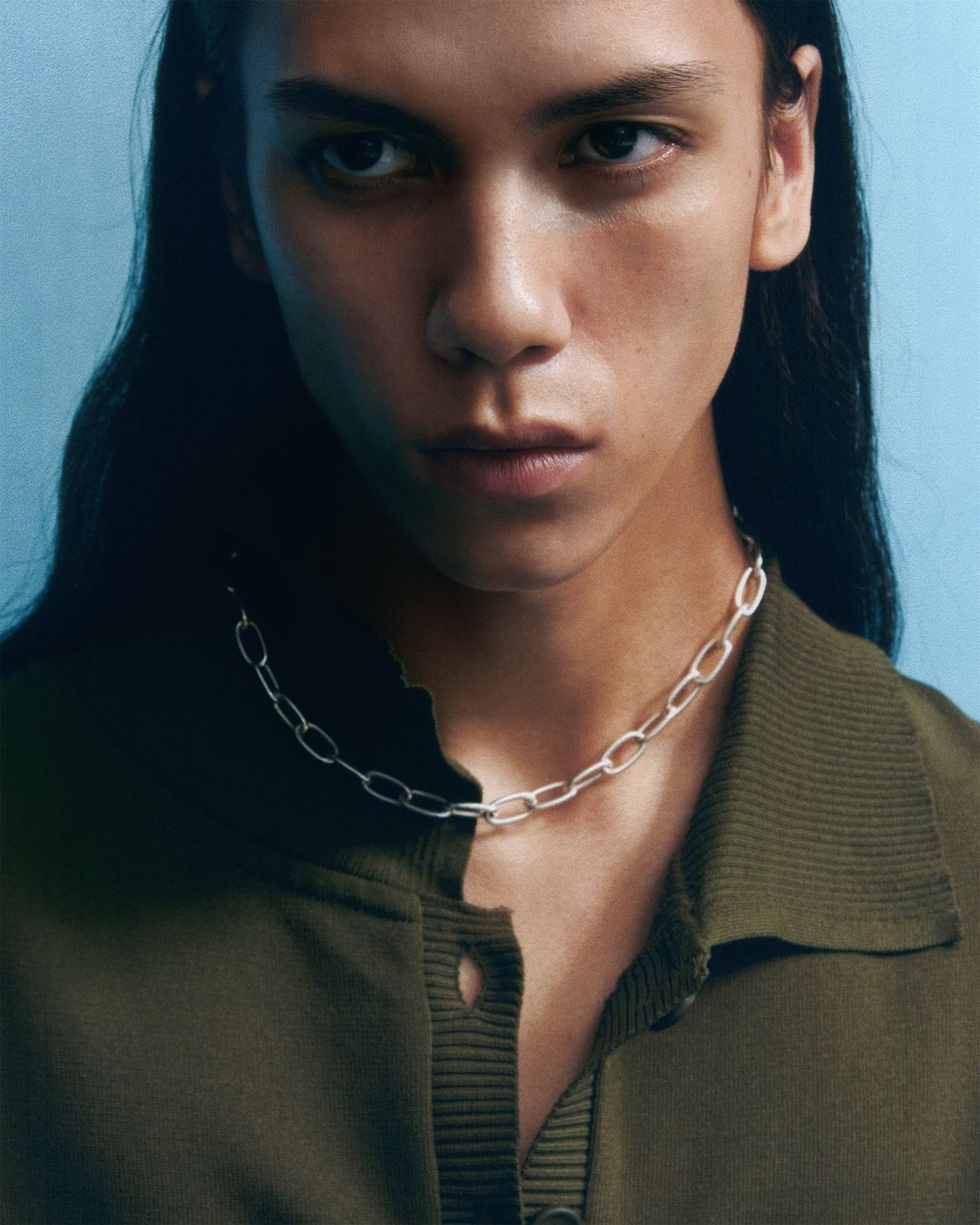 A young adult with long dark hair models a silver chain necklace, wearing an olive green collared shirt against a light blue background.