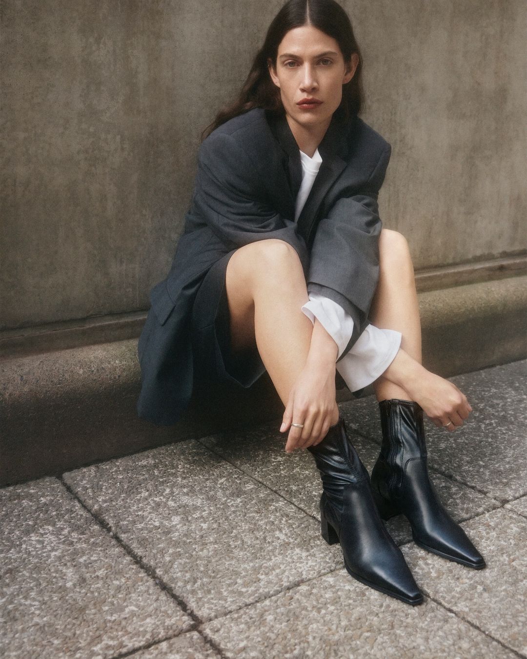A woman with long brown hair sits on stone steps wearing a grey blazer, black shorts, a white shirt, and black leather boots.