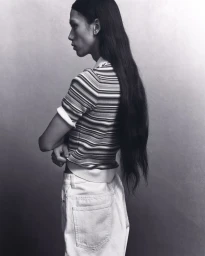 Model with long dark hair wearing a striped polo shirt and white trousers against a neutral wall by Ellen Nykvist.