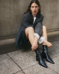 A woman in an oversized grey blazer and black boots sitting on stone paving against a concrete wall, photographed in Stockholm.