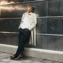 Man leaning against a dark metallic wall wearing a beige trench coat, white shirt, dark trousers, and brown leather boots.