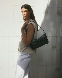 Model in sleeveless cream top and white trousers posing outdoors against a pale textured tile wall carrying a black shoulder bag
