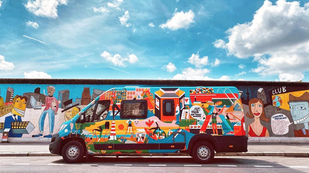 Art-covered camper van parked on a city street in front of mural art wall on a sunny day with blue skies and wispy clouds.