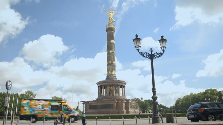 Victory Column in Berlin, Germany under a partly cloudy sky, viewed from street level with traffic and greenery in the background.
