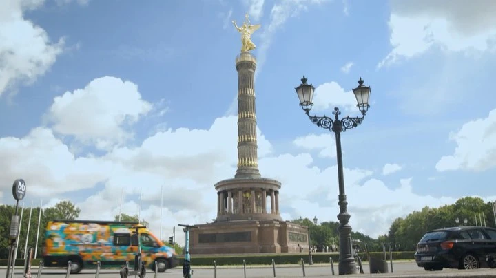 Victory Column in Berlin, Germany under a partly cloudy sky, viewed from street level with traffic and greenery in the background.